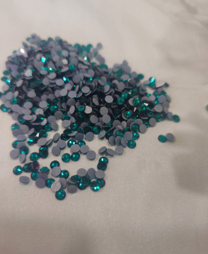 Teal and green flatback rhinestones are scattered on fabric backgrounds, with green leaves nearby.