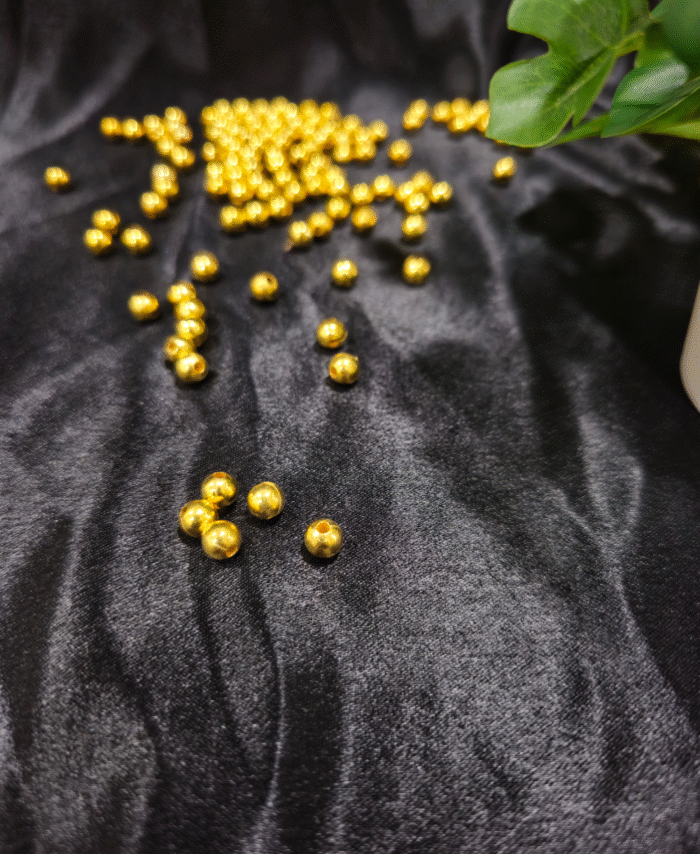 Golden pearl beads are scattered on a black silky fabric with green plant leaves in the corner.