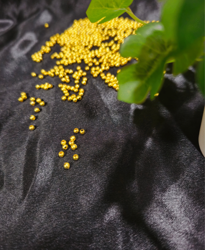 Golden pearl beads are scattered on a black silky fabric with green plant leaves in the corner.