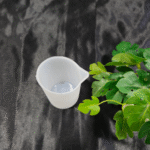 Transparent plastic measuring cup placed on dark background with green plant.