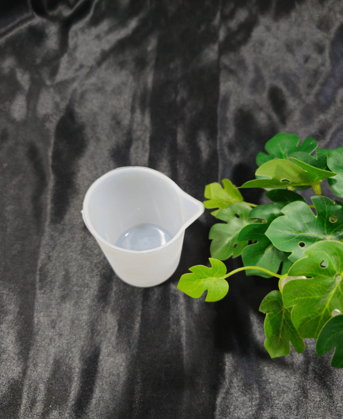 Transparent plastic measuring cup placed on dark background with green plant.