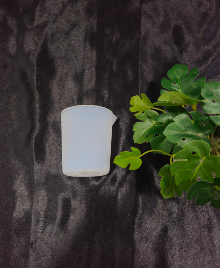 Transparent plastic measuring cup placed on dark background with green plant.