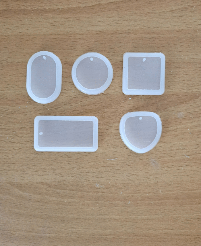 Five silicone keychain moulds in oval, round, square, rectangular, and shield shapes are placed on a wooden surface. Transparent silicone moulds for resin keychain making in assorted shapes.