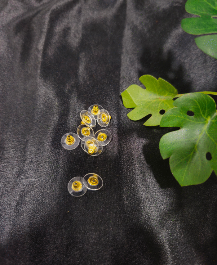 Clear flat earring backs with gold centres placed on a black fabric background with green leaves beside them.”