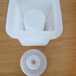 Square silicone dispenser mould with a central pillar and a separate round lid insert placed on a wooden surface White silicone mould for making resin or soap dispensers with a detachable circular top piece