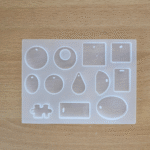 Transparent multi-shape silicone earring mould with round, oval, square, teardrop and puzzle shapes placed on a wooden surface.
