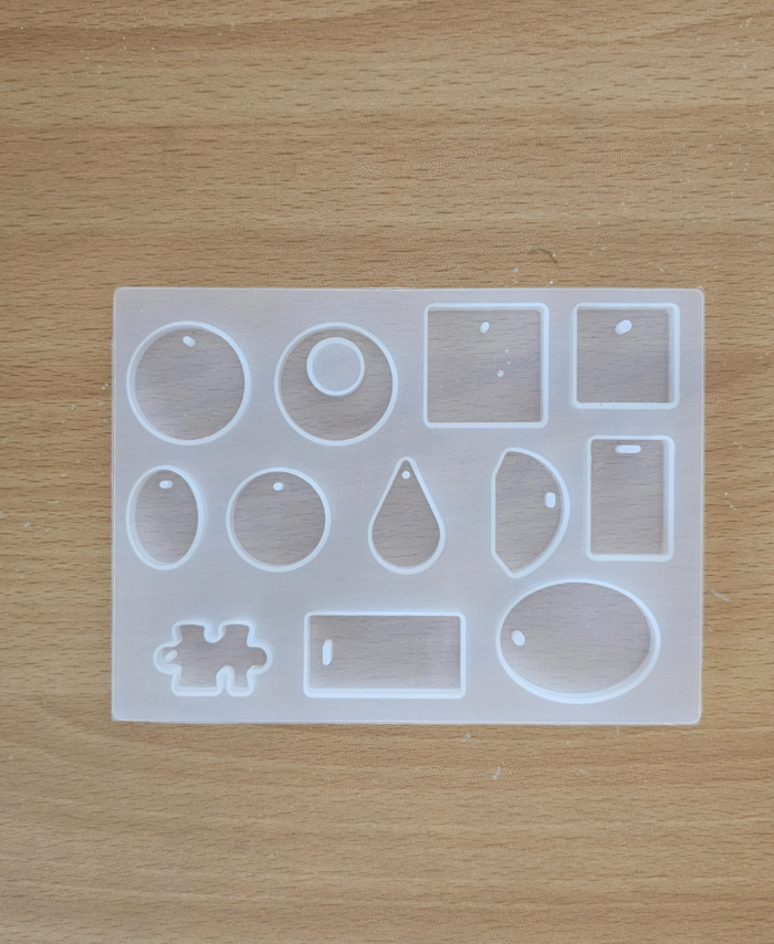 Transparent multi-shape silicone earring mould with round, oval, square, teardrop and puzzle shapes placed on a wooden surface.