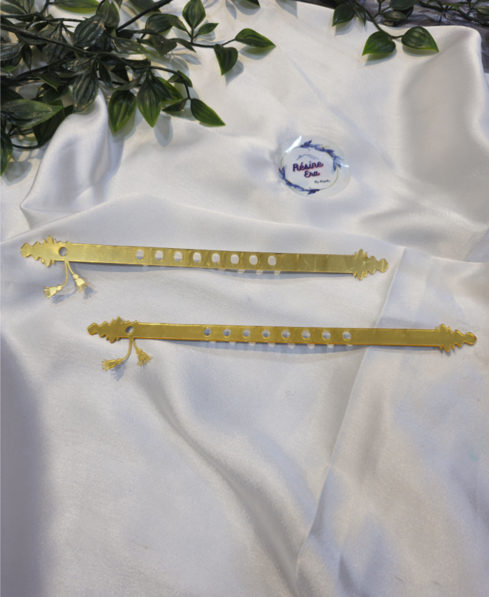 Two golden acrylic basuri strips with tassels, decorative ends, and circular holes are displayed on white fabric.