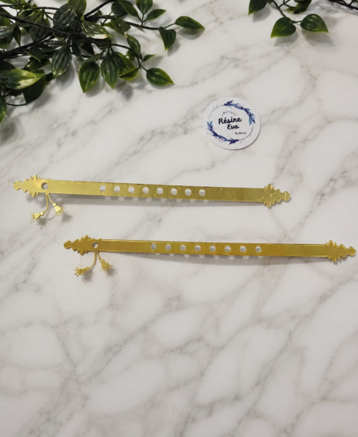 Two golden acrylic basuri strips with tassels, decorative ends, and circular holes are displayed on white fabric.