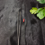 A slim precision tweezer tool is placed on a black background with green leaves beside it.