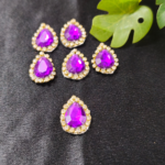purple teardrop embellishment stones with a diamond border are placed on a black background with green leaves beside them.