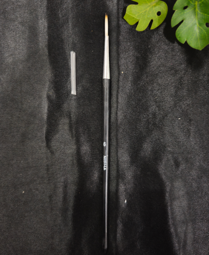 Thin resin art detail brush with protective cap placed on black background near green leaves.