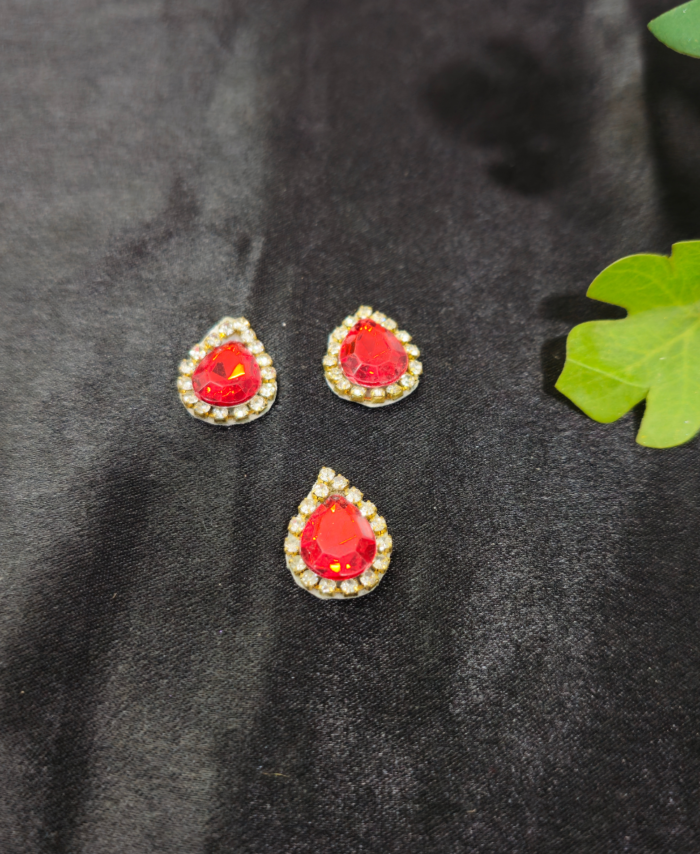 Red teardrop embellishment stones with a diamond border are placed on a black background with green leaves beside them.