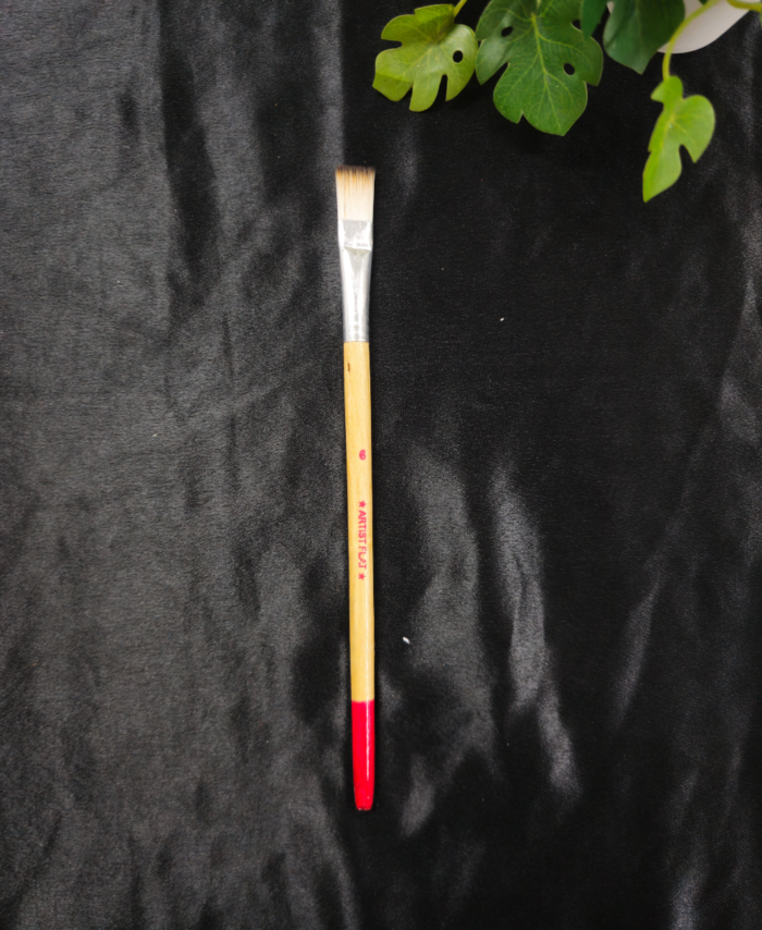 Thick resin art flat brush with protective cap placed on black background near green leaves.