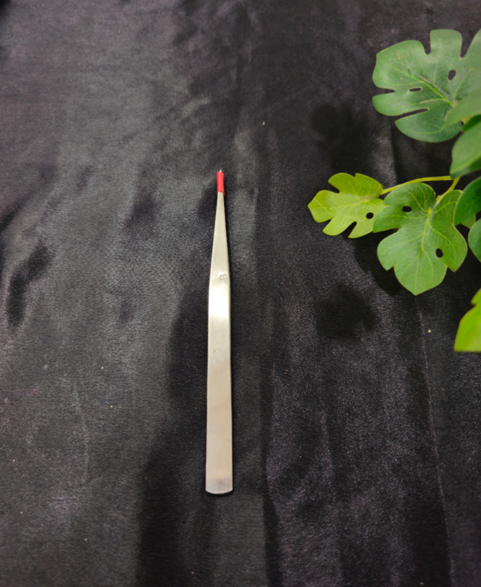 A slim precision tweezer tool is placed on a black background with green leaves beside it.